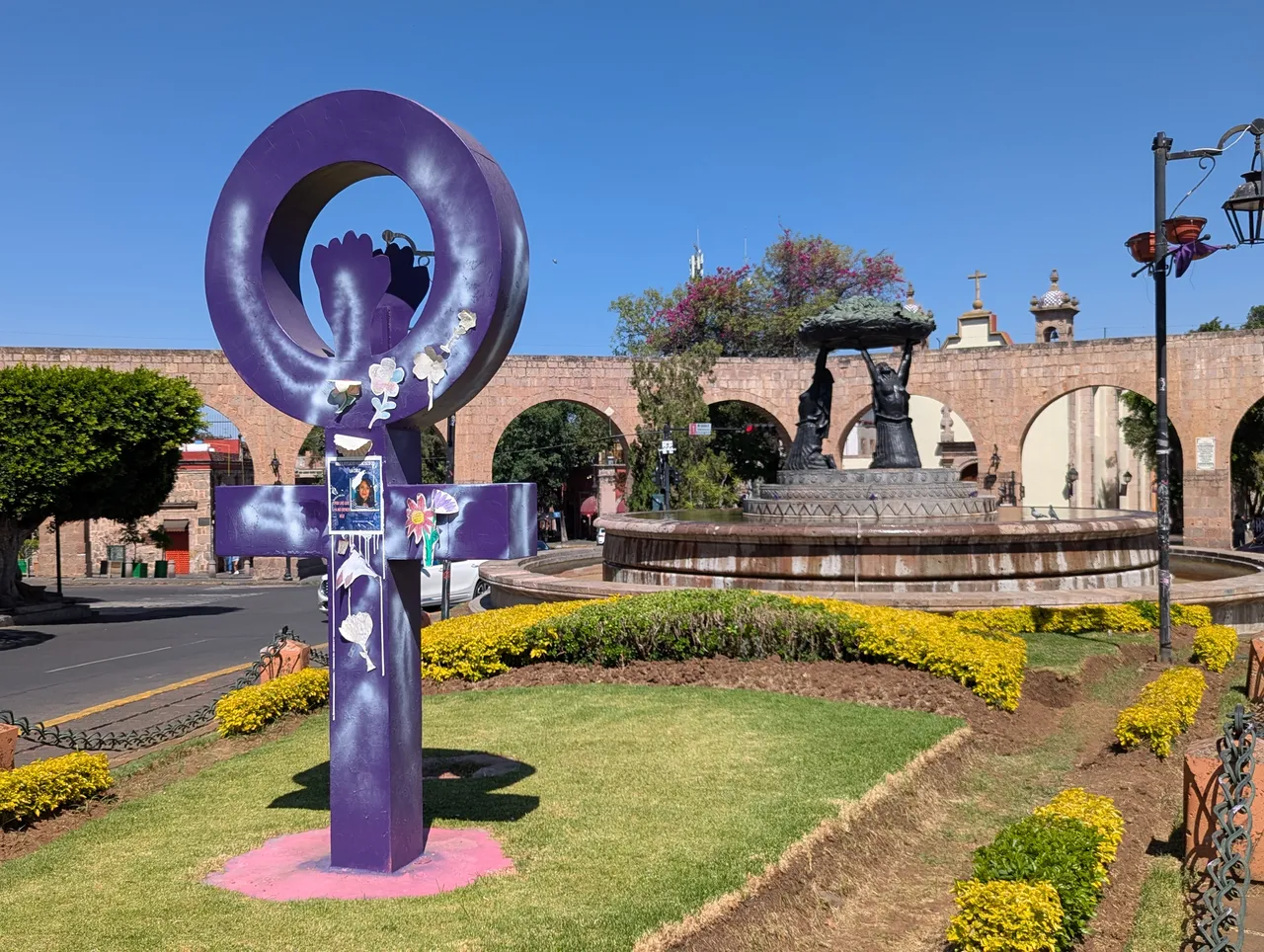 Lila Skulptur und Brunnen neben Morelias Stein-Aquädukt in Mexiko, mit entfernten Fußgängern bei hellem Sonnenschein