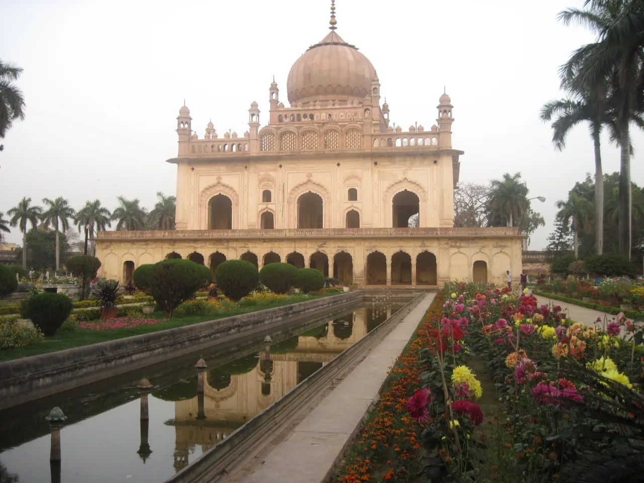 The historic Gulab Bari mausoleum in Faizabad, India, featuring Mughal architecture, a reflective water canal, manicured gardens, and visitors under soft daylight.