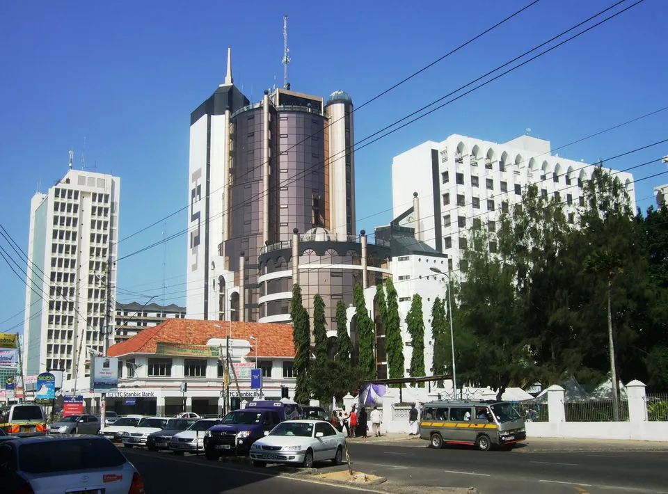 A sunny street scene in Mombasa, Kenya, featuring modern high-rise glass buildings, a colonial-style bank, and local traffic on a clear blue day.