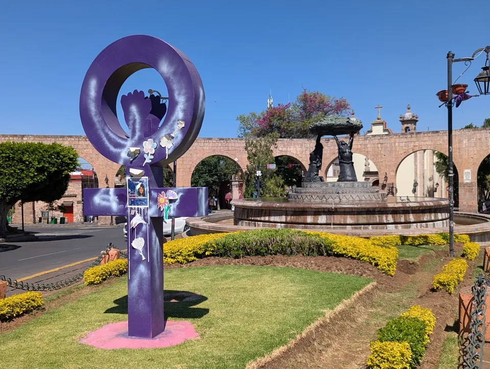 Escultura y fuente moradas junto al acueducto de piedra de Morelia en México, con peatones distantes bajo un sol brillante
