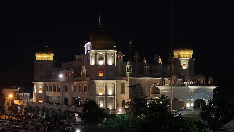 A stunning night view of a brightly illuminated Gurdwara in Ludhiana, India, featuring golden domes, intricate white architecture, and surrounding trees.